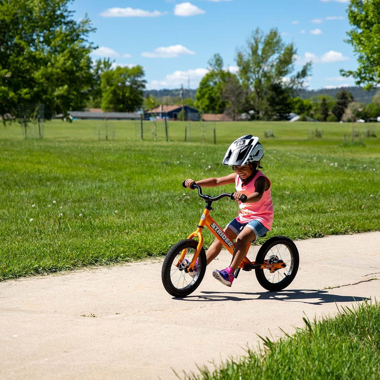 Child riding a balance bike on a paved path with green grass and trees in the background