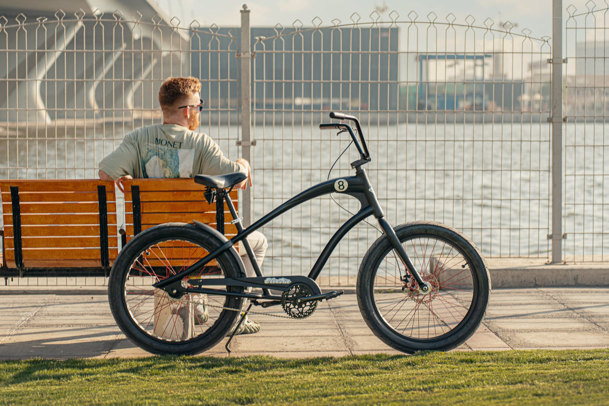 Man sitting on a bench with a black bicycle next to him, against a metal fence and building background.