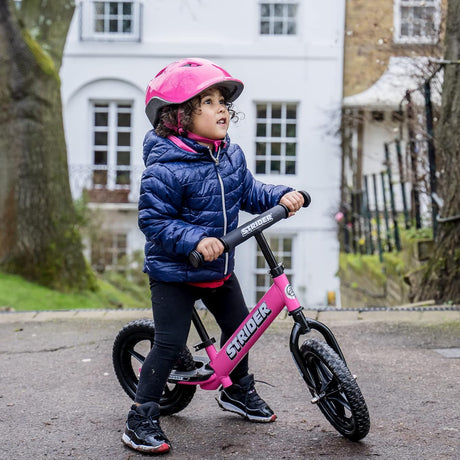 Child wearing a pink helmet and blue jacket riding a pink Strider balance bike on a residential street.